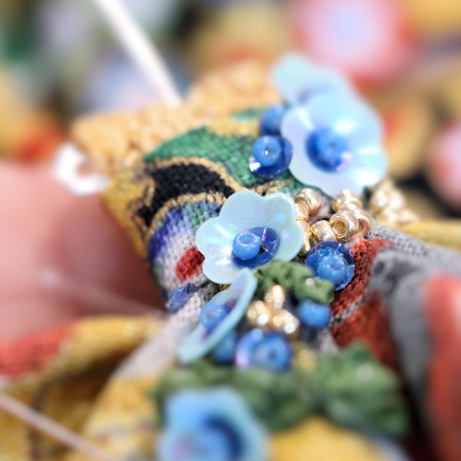 Zoom sur la broderie d'art miniature d'une éclosion de fleurs en ruban de soie sur la robe d'un pendentif poupée talisman. Murmures d'une charmeuse